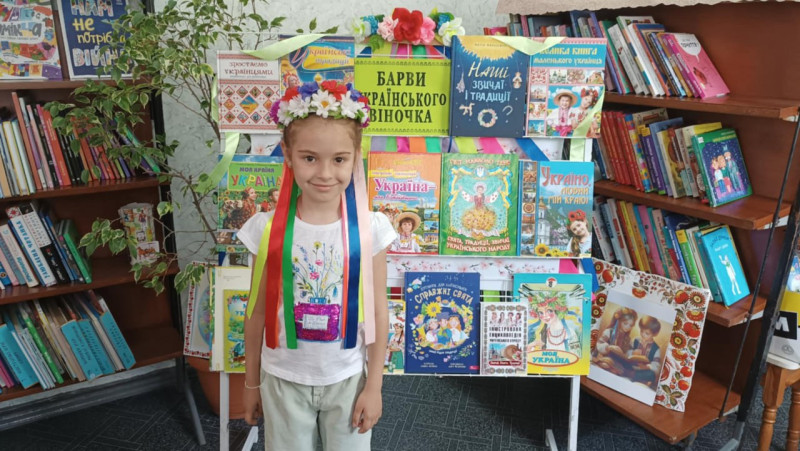 Дівчинка у віночку стоїть перед стендом з книжками, об'єднаними загальною назвою "Барви українського віночка". Навколо - полиці з книгами.