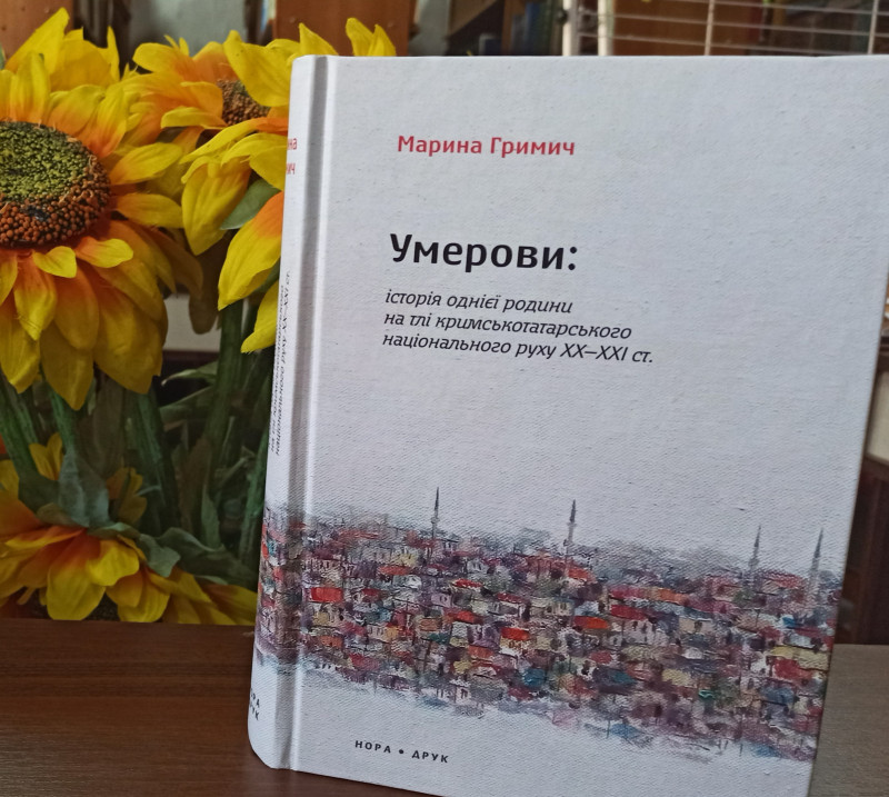 На столі стоїть книга  Марини Гримич "Умерови: історія однієї родини на тлі кримськотатарського національного руху XX -XXI ст." За книгою  - букет із соняшників.