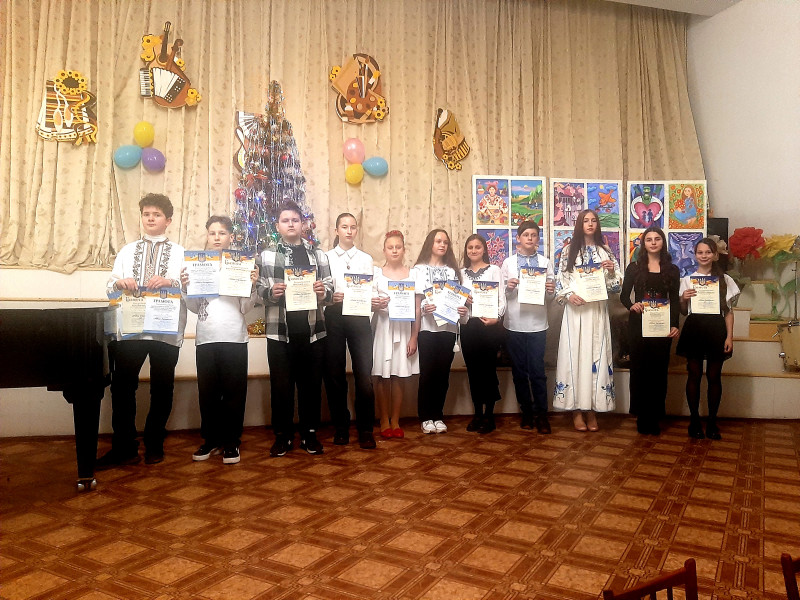 Дівчата та хлопці стоять на сцені, в руках тримають грамоти. На задньому плані  - прикрашена ялинка, кольорові кульки, виставка робіт художників,  емблеми відділів школи. 