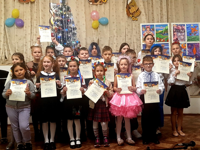 Діти стоять  гуртом, тримаючи в руках грамоти. На задньому плані  - прикрашена ялинка, кольорові кульки, виставка робіт художників, емблеми відділів школи.                                            