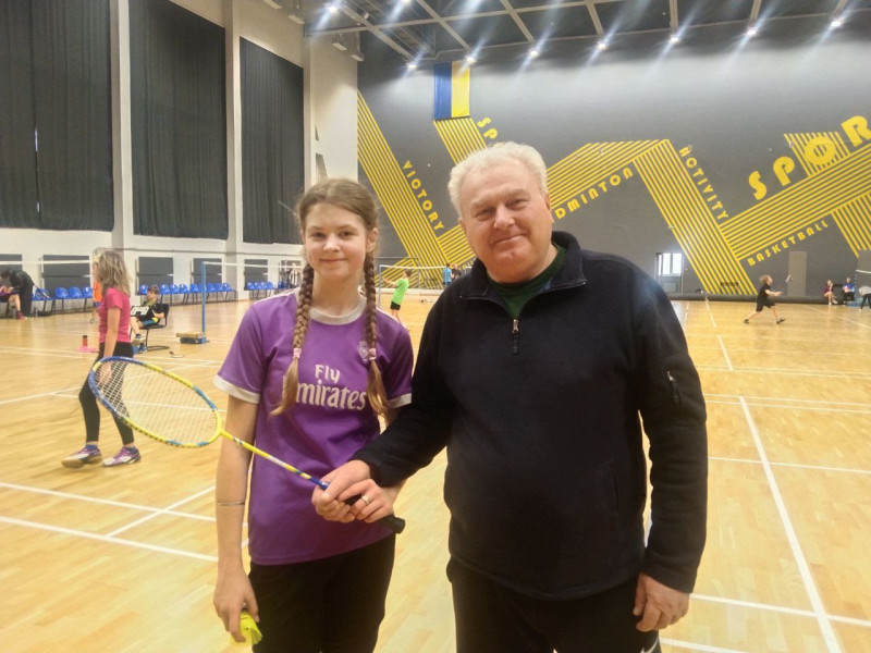 У спортивній залі стоїть тренер з ракеткою, поряд - усміхнена дівчинка з воланчиком в руці. 