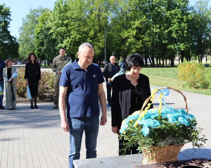Міський голова Дмитро Зражевський та начальник Синельниківської районної військової адміністрації Світлана Назарчук стоять, схиливши голови перед кошиком із квітами. За ними  - інші представники міської та районнної влади, військовий із букетами квітів.