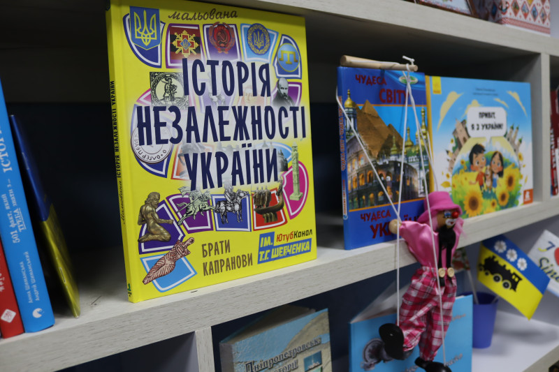 На полицях стоять книги. На передньому плані книга під назвою "Історія незалежності України".