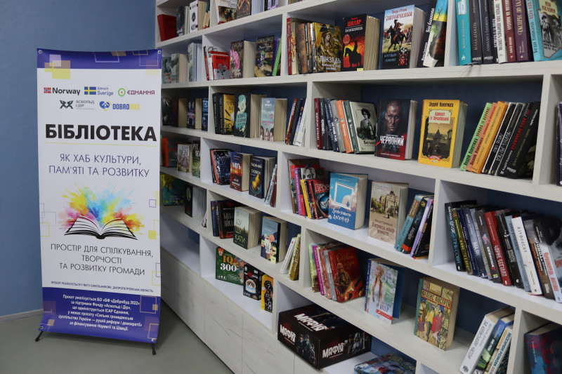 На полицях стоять книги. Поряд  - банер із назвою проєкта, учасниками. 