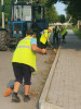 Жінки у жилетках  прибирають мітлами вздовж пішохідної доріжки. За ними - трактор.
