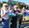 Жінки, різні за віком, стоять з букетами блакитно-жовтих квітів, інші тримають в руках  Прапор України. 