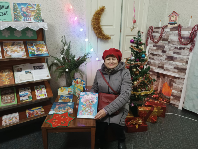 Хол у бібліотеці. Тут - поличка і стіл із книжками новорічної тематики. Відвідувачка закладу тримає одну із книг у руках, усміхаючись. За нею - прикрашена ялинка, під якою - коробки -подарунки.  