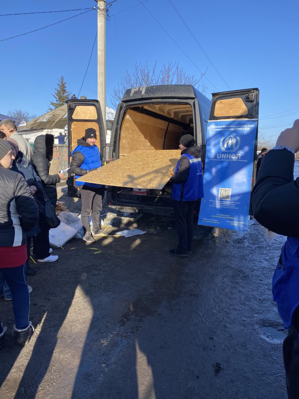 Представники "Проліски" - два чоловіки тримають в руках лист ОСБ плити, щоб передати людям, які стоять в черзі. Лист дістали з  машини.