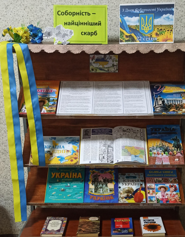 Книжкова полиця з літературою. З верхнього лівого кутка полиці  звисають жовто-блакитні стрічки, тут же розташовані маленькі сині та жовті квіточки, карта України. Праворуч на верхній полиці таблички з написами: "Соборність - найцінніший скарб" та "22 січня. З Днем Соборності України!"