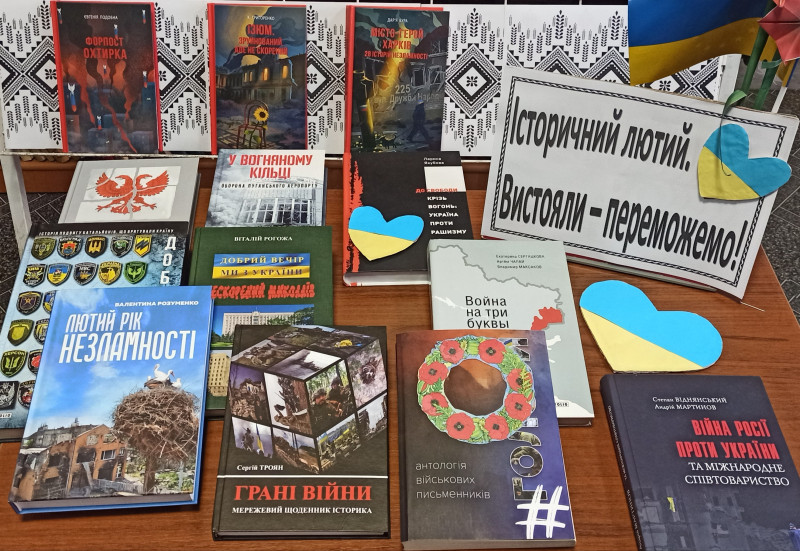 на столі  -книжкова виставка під назвою "Історичний лютий. Вистояли - переможемо!". Між книгами - синьо-жовті сердечки. В правому верхньому кутку - Прапор України. 