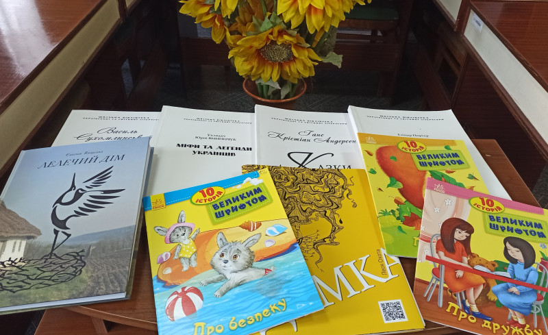 На столі лежать книги, серед них  із назвами: "10 історій великим шрифтом. Про безпеку", "10 історій великим шрифтом. Про дружбу". За столом на підлозі - квіти соняха у вазоні..
