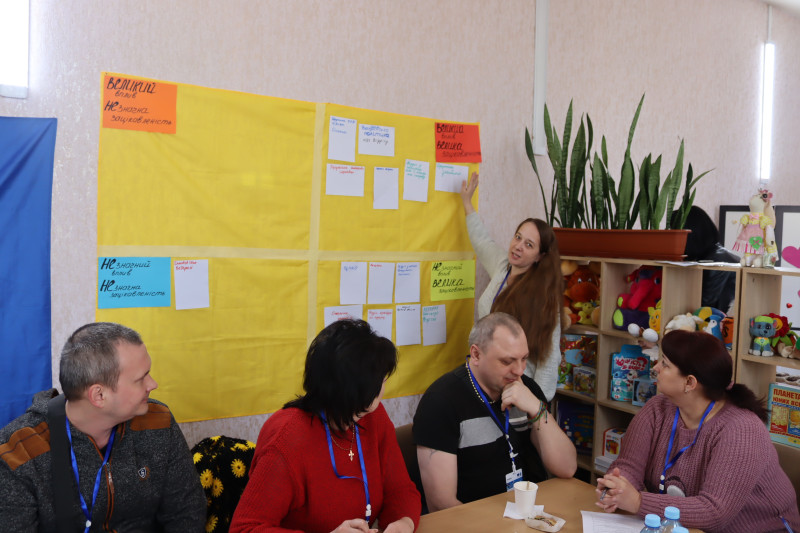 За столом сидять учасники заходу. Одна із учасниць стоїть біля жовтого полотна, на яке розмістила свою листівку, підтримує її рукою.