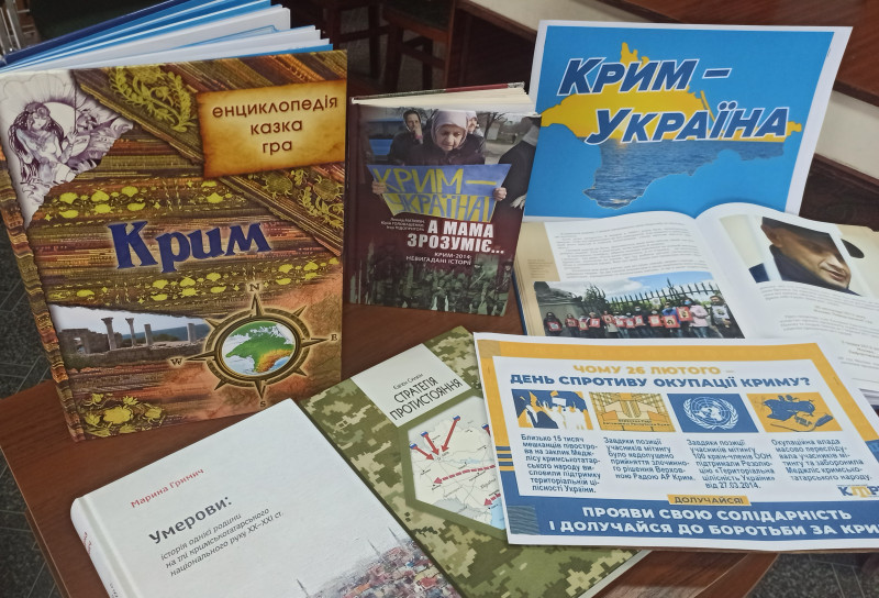 На столі - книги, кілька стоять, інші - лежать. На столі також знаходиться  листівка  з написом:  "Крим - Україна". 