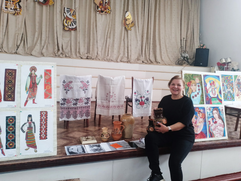 Викладач школи Світлана Вовк з глечиком в руках.  Поряд стоять інші.  На стільцях висять вишиті рушники. Стоять стенди з зображеннями українок в різних костюмах та окремими елементами вишитих візерунків. На інших стендах - картини з українками, намальовані учнями школи. 