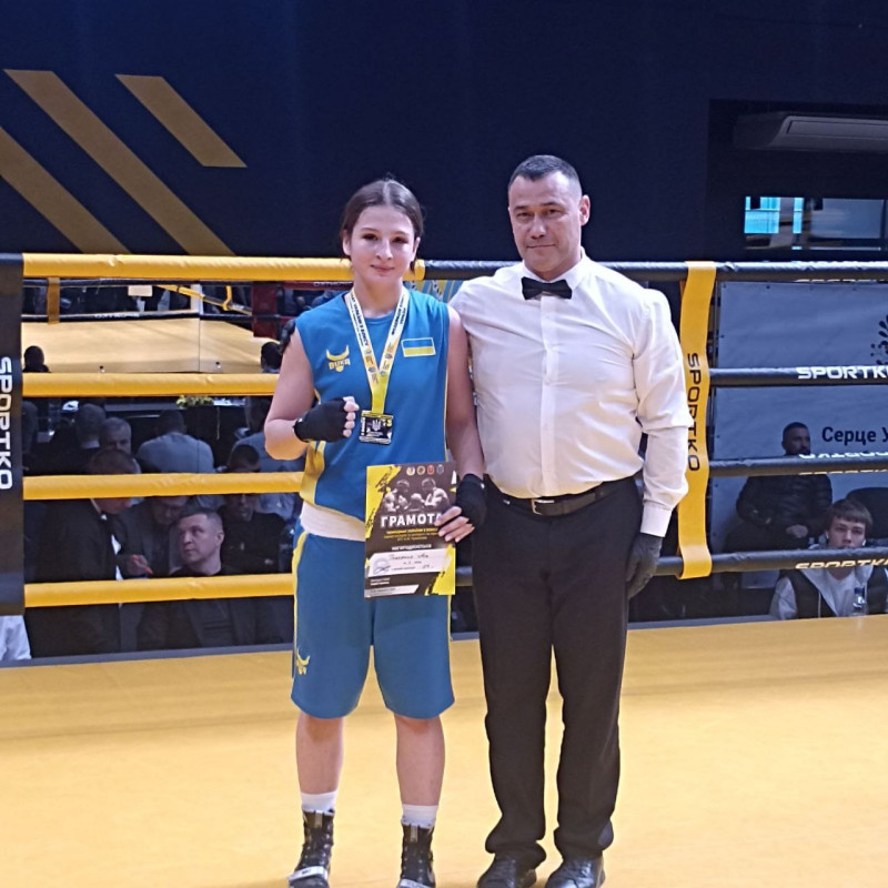 На боксерському ринці стоїть боксерка Ася Ткаченко з суддею. У руках дівчина тримає грамоту, на шиї -  медаль. 