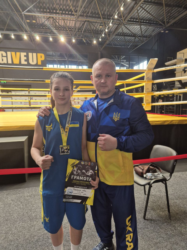 Біля боксерського рингу стоїть боксерка Ася Ткаченко з тренером Максимом Гончаром. У руках дівчини -  грамота, на шиї -  медаль. По одній руці  зігнули в ліктях.