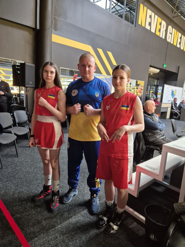 У спортивній залі тренер Максим Гончар стоїть разом із вихованками - Ткаченко Асею та Дроновою Богданою.  Руки - зігнуті в локтях.