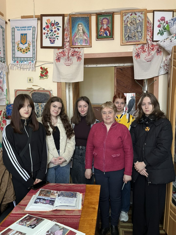 Керівник клубу "Муліне" Людмила Савченко стоїть в оточенні дівчат. Над ними спускаються вишиті рушники, висят вишиті картини. На столі - розкриті альбоми з фото.