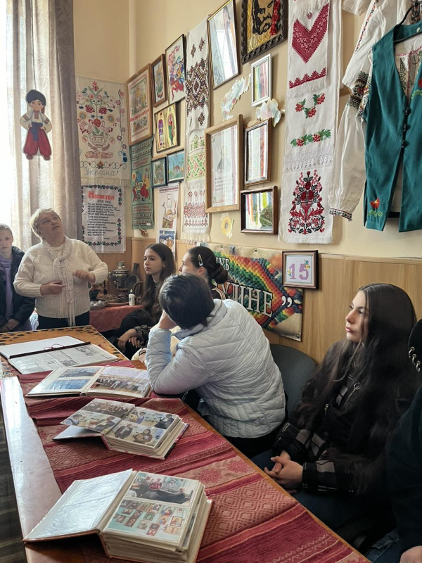 Керівник клубу "Муліне" Людмила Савченко розповідає дітям про експонати виставки -  вишиті рушники, картини, що висять на стіні. Слухачі сидять за столом, інші - стоять  поруч.  На столі лежать альбоми з фото.