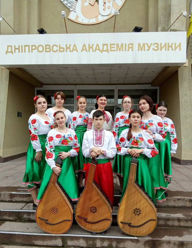 Група людей стоїть на сходах до Дніпровської академії музики, серед яких: дівчата, юнак, викладачка, всі  в українських костюмах. Дві дівчини та юнак у першому ряду сперлись на бандури.  