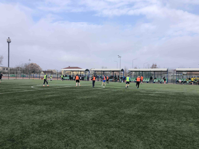 Футболісти під час гри на полі. Вдалині - секції зі стільцями та навісами. Тут сидять вболівальники.