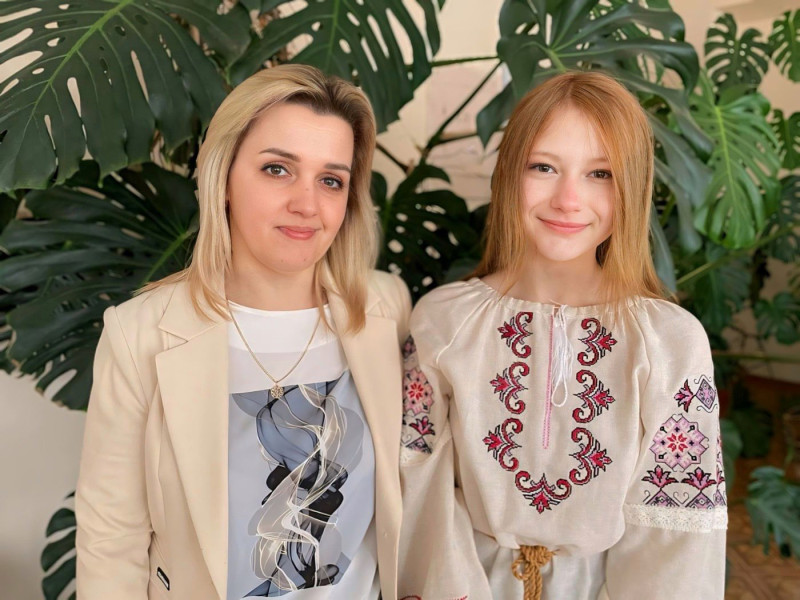 Вчителька разом зі своєю ученицею стоять на фоні зелені - рослини монстери.