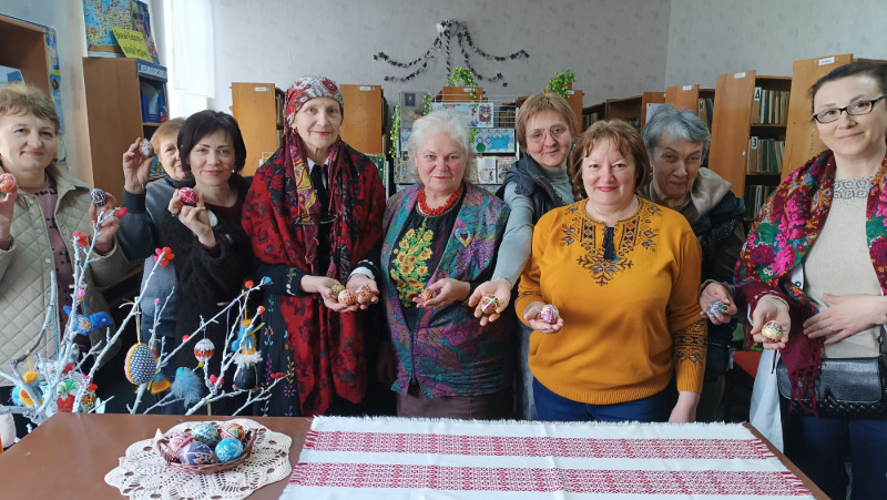 Учасниці заходу  - жінки, різні за віком, усміхнені, тримають у руках писанки. Перед ними - стіл  з вишитою скатертиною, серветкою, на якій - маленький кошик з писанками.  За жінками - стелажі з книгами.