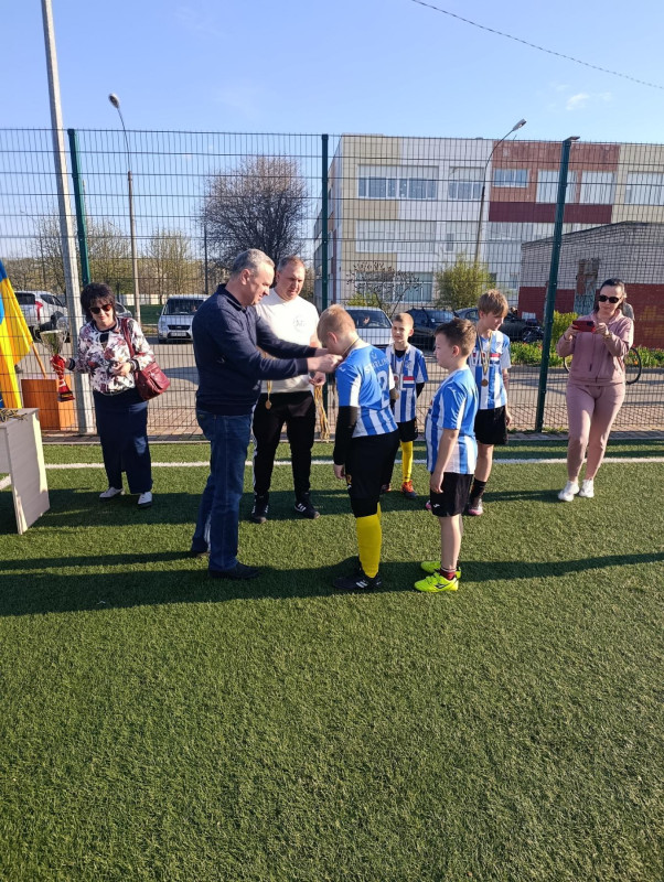 Міський голова надягає медаль юному футболісту. За цим футболістом стоїть інший.  На задньому плані - матусі, організатори турніру.