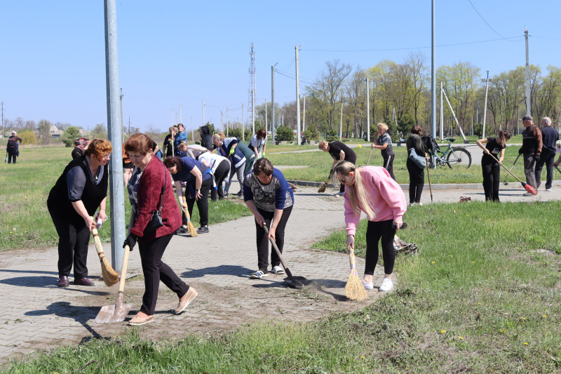 На передньому плані - група з  чотирьох жінок за роботою на доріжці: хтось з лопатою, хтось із виником.  За ними - інші люди теж працюють. 