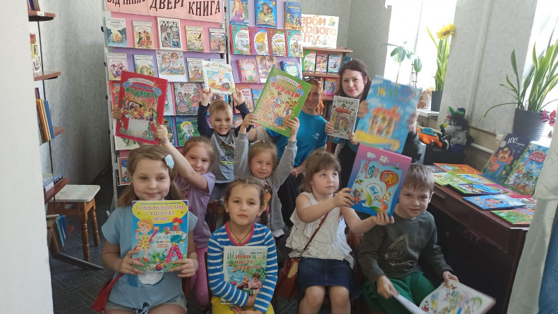 Усміхнені діти  із книжками в руках разом із бібліотекарем  біля стенду з книгами