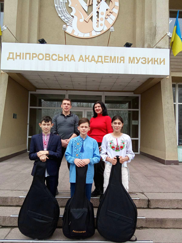 Група людей з п'яти осіб: викладач, викладачка, два хлопця і дівчина стоять на сходах до Дніпровської академії музики. Двоє з дітей сперлись на бандури в чорних чохлах , один хлопець - на гітару  в чорному чохлі.