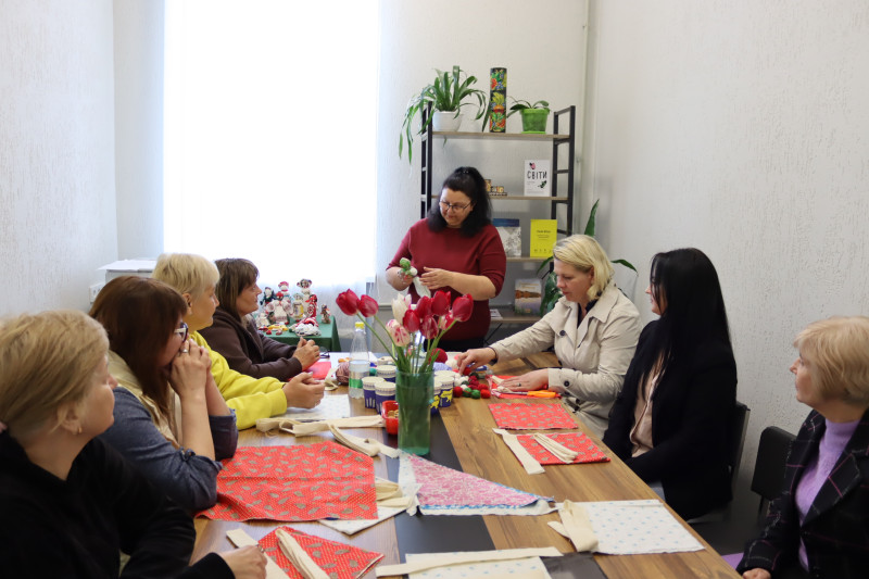 Жінки сидять за столом, на якому лежить тканина, нитки, стоять квіти у вазоні. Майстриня показує та розказує, що треба робити.