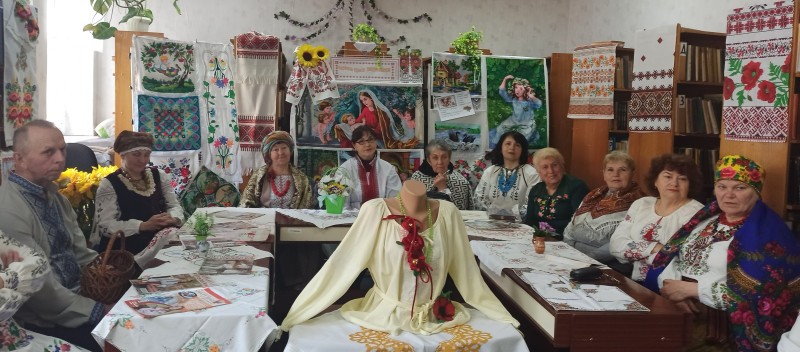 За столами у залі бібліотеці сидять жінки в українському вбранні. Є і один чоловік. На столах - журнали, вишивки. На полицях із книгами - вишиті картини, рушники.  Посередині - невеликий стіл із манекеном, на якому - жіноча сорочка та  прикраса з червоних квітів.