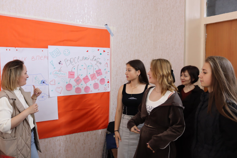 Група дівчат біля дошки. Одна пояснює, що намальовано на плакаті з написом: "Веселий  Хабрик".