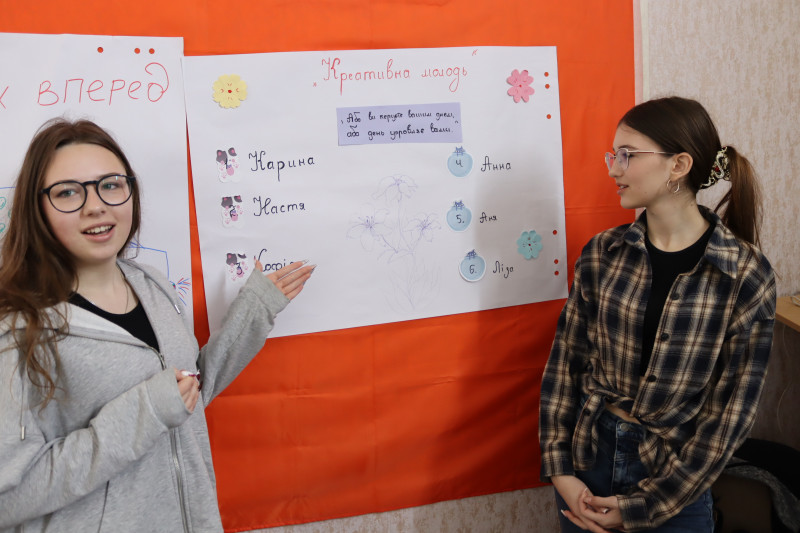 Дві дівчини знаходяться біля дошки з плакатами. Одна з них пояснює, що намальовано на плакаті з написом: "Креативна молодь".