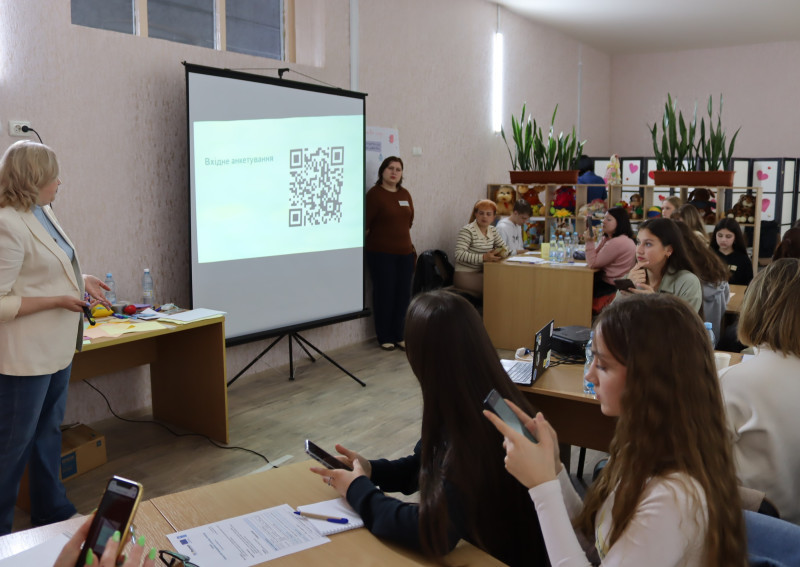 Учасники сидять за столами. У руках тримають телефони, проходять вхідне тестування. Біля стіни стоїть екран з QR-кодом.   