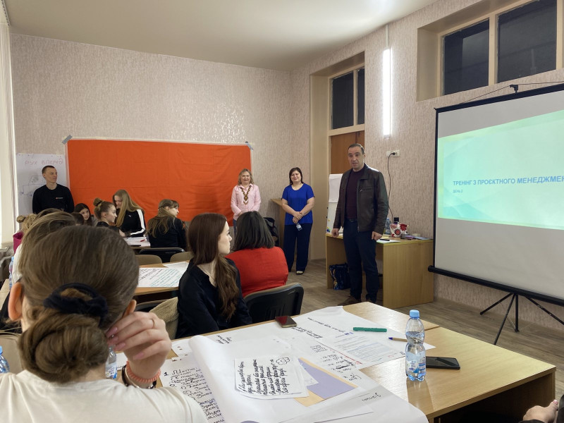 Учасниці заходу сидять за столами. Перед ними -  спікери - дві жінки та чоловік. На екрані слайд із назвою тренінгу.