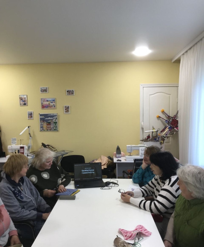 За столом сидять жінки. Погляди направлені на монітор ноутбука, де на екрані напис: "Безбар'єрна грамотність".