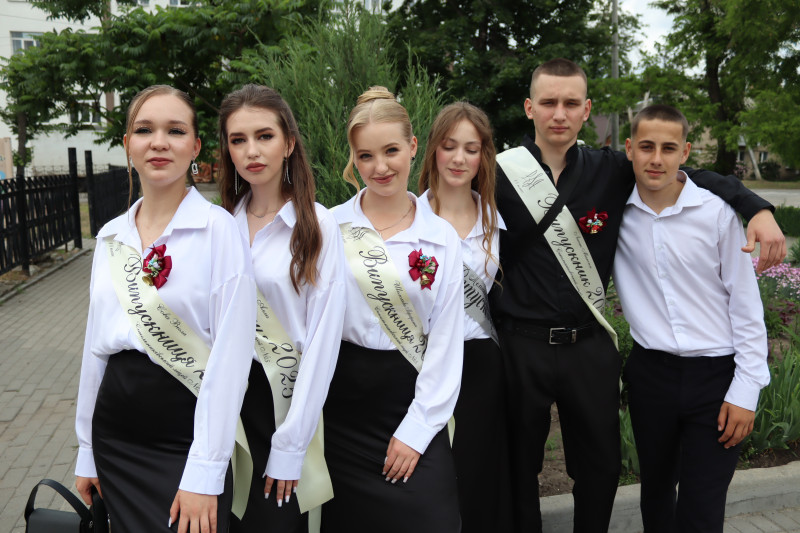 Група ліцеїстів з білими стрічками через плече,  маленькими дзвіночками  на грудях  стоїть на фоні зелені.