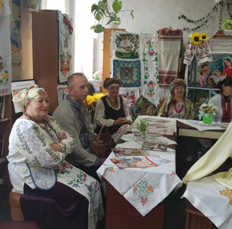 За столом сидять жінки та чоловік в українському вбранні. На столі - журнали, вишиті серветки. З книжкових полиць звисають вишиті рушники. 