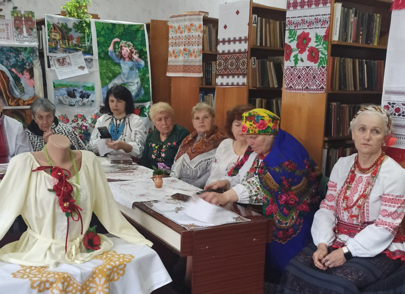 За столом сидять жінки в українському вбранні. На столі - вишиті серветки. З книжкових полиць звисають вишиті рушники. Висять вишиті картини. На окремому столі стоїть манекен. На ньому  - сорочка, намисто з червоними квітами.      