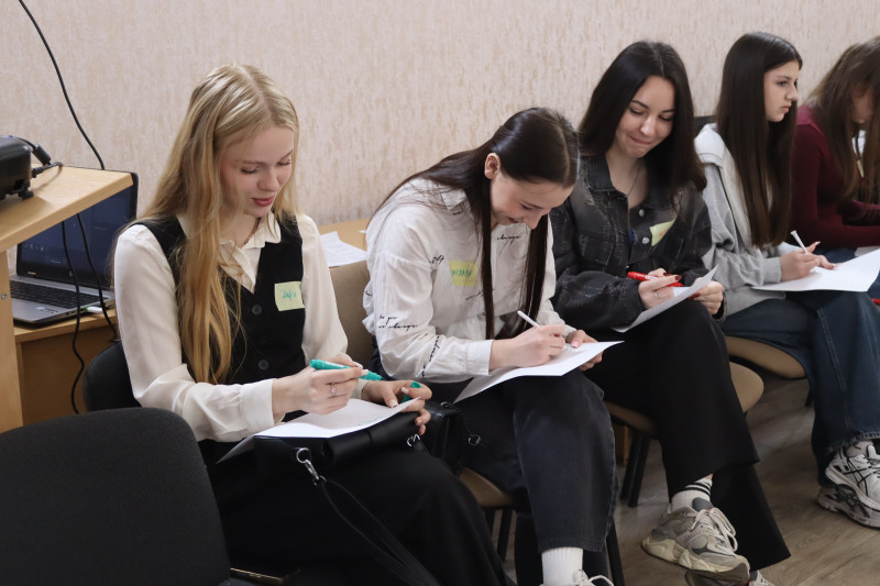 Чотири дівчини сидять на стільцях. Кожна щось пише фломастером  на папері, усміхаючись.