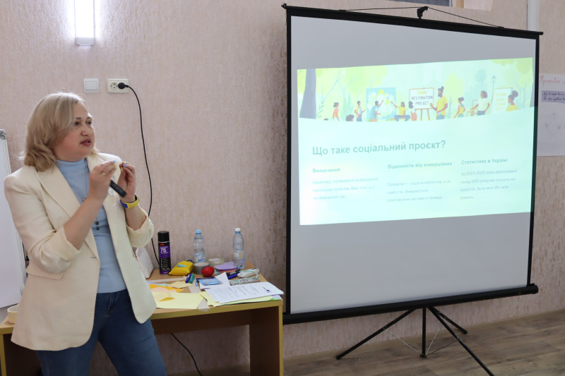 Тренерка стоїть біля екрану і розповідає. На екрані - слайд, де напис: "Що таке соціальний проєкт?"  та інші.