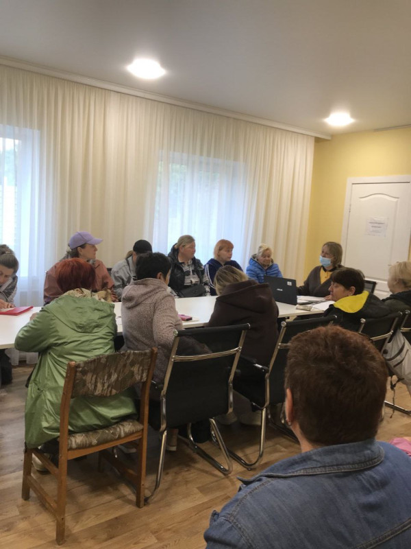 За довгим столом сидять жінки. Погляди спрямовані на жінку в медичній масці на обличчі, що сидить у торці столу.  Перед жінкою - відкритий ноутбук.