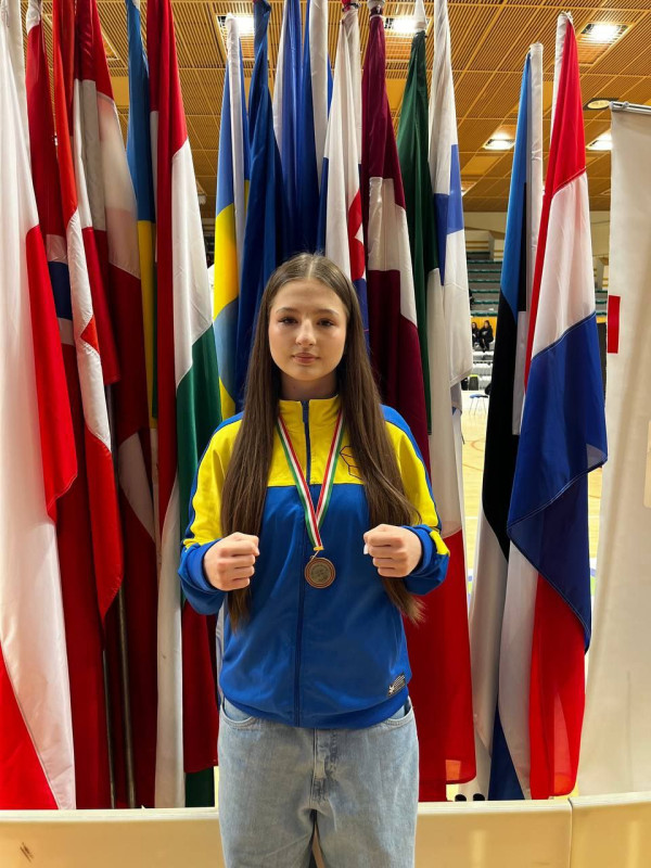 Спортсменка у кофті з синьо-жовтими кольорами  стоїть на фоні різних прапорів, зігнувши пальці в кулаки.  На грудях - медаль.