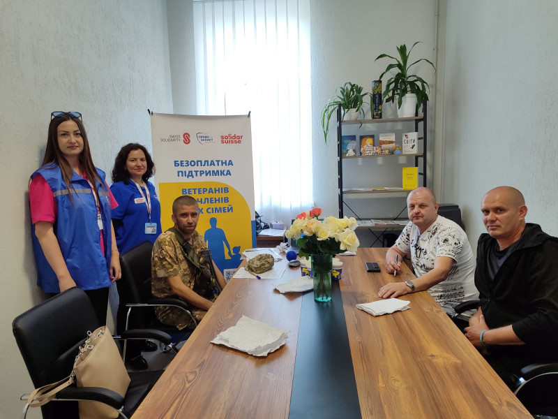 У кімнаті за столом сидять три чоловіка, один - у військовій формі.  За ними стоять дві жінки, стенд з емблемами та написом: "Безоплатна підтримка ветеранів та членів їх сімей", полиця.