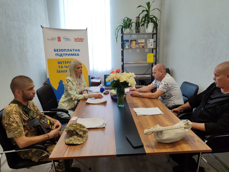 У кімнаті за столом сидять три чоловіка, один - у військовій формі, і жінка. За ними -  стенд з емблемами та написом: "Безоплатна підтримка ветеранів та членів їх сімей", полиця.