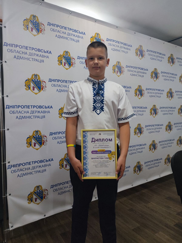 На фоні банеру Дніпропетровської обласної державної адміністрації стоїть хлопчик у вишиванці та тримає в руках диплом