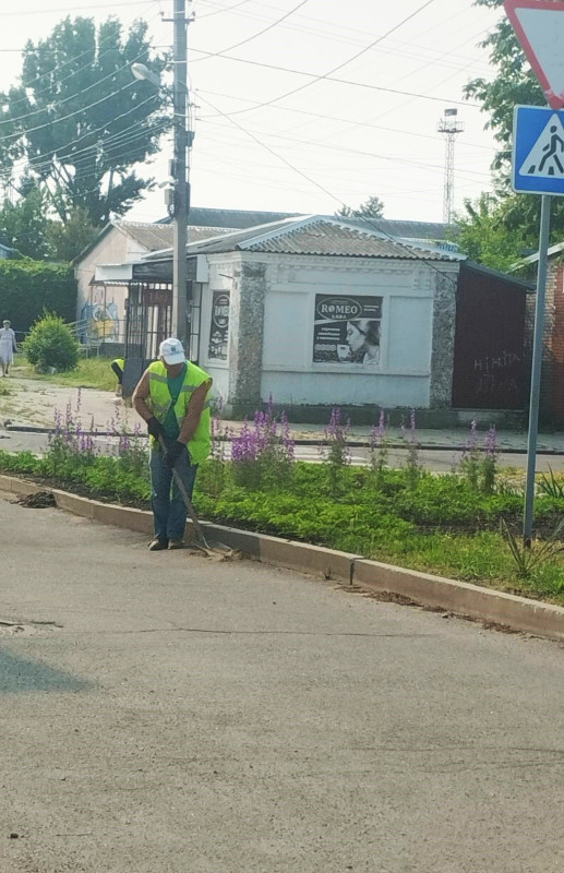 Чоловік у жилетці салатного кольору  прибирає лопатою вздовж бордюра.