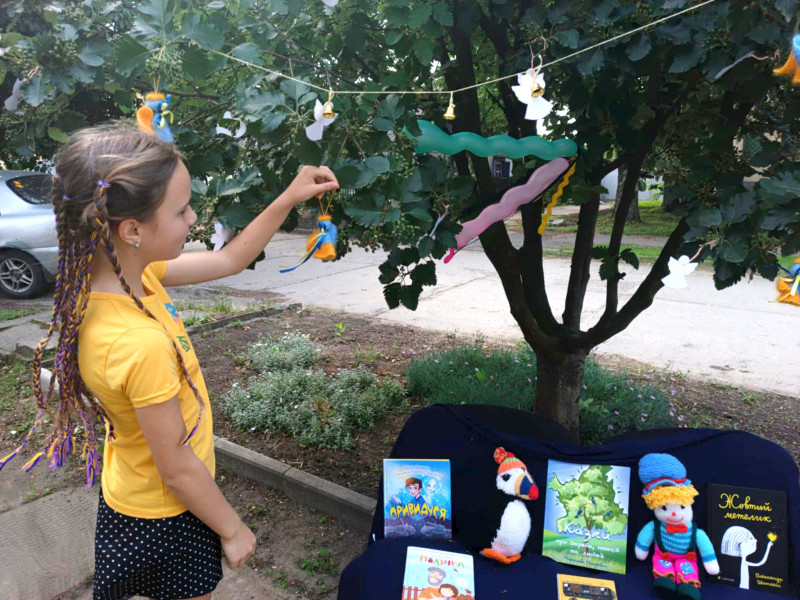 Дівчинка чіпляє янгола на дерево. Під деревом - лава з книжками, дитячими іграшками.